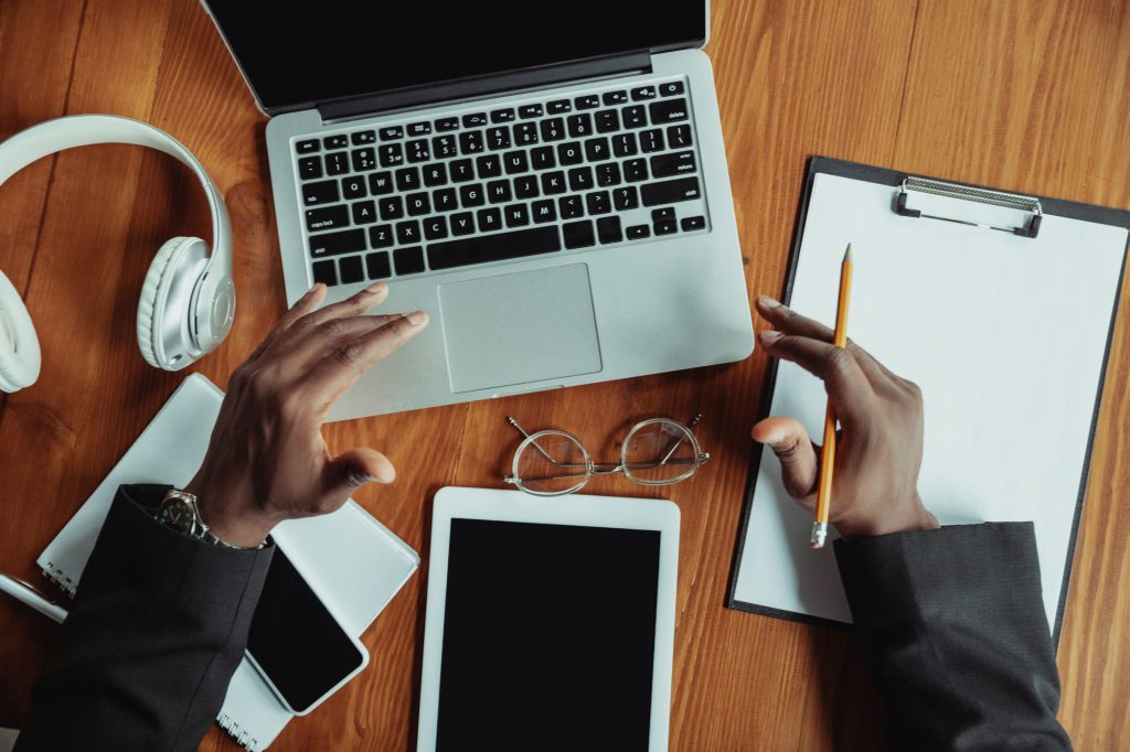 Black man hands over a laptop , a tablet , a phone , a clipboard , glasses and headphones on a wooden desk for simple web3 content creation.